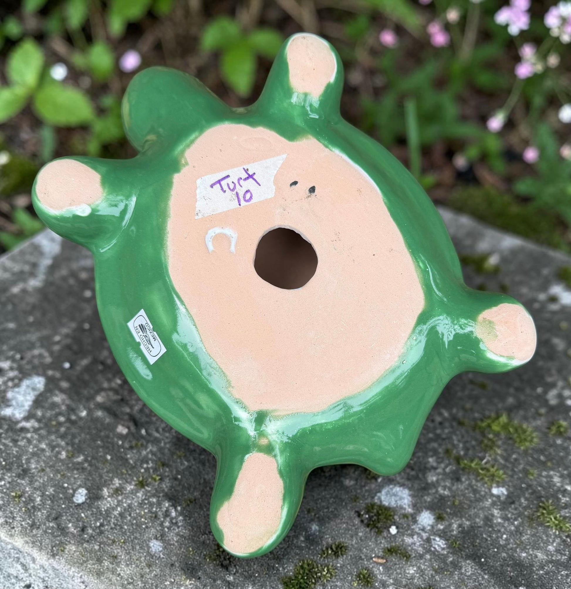 A hand-painted ceramic turtle figurine resting on a gray stone surface with a blurred green garden background. The turtle features a bright green head and legs, and a highly detailed, multicolored shell with yellow, red, orange, blue, and black patterns, bottom view of turtle,