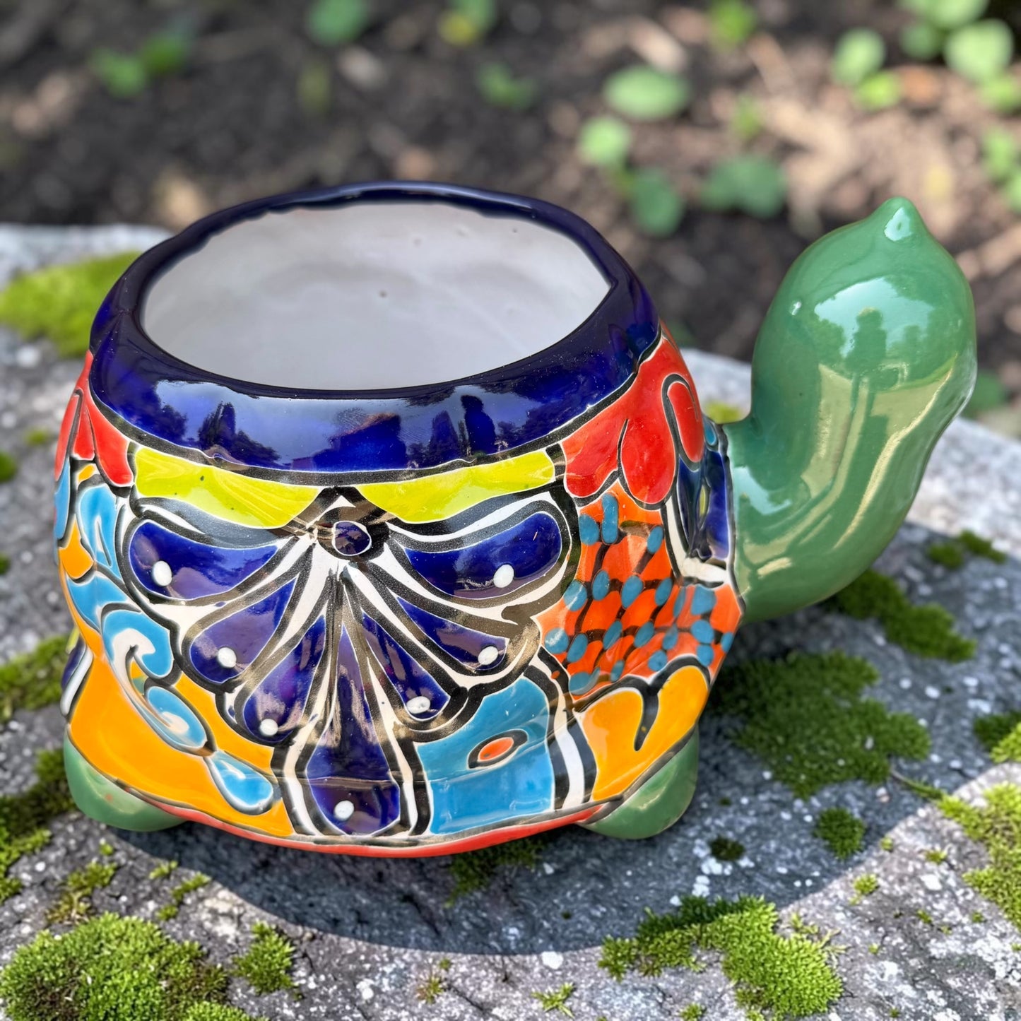 A colorful ceramic turtle-shaped planter with a green head and legs, decorated with vibrant hand-painted patterns in orange, yellow, blue, red, and white, sitting on a mossy stone outdoors. Back of turtle.
