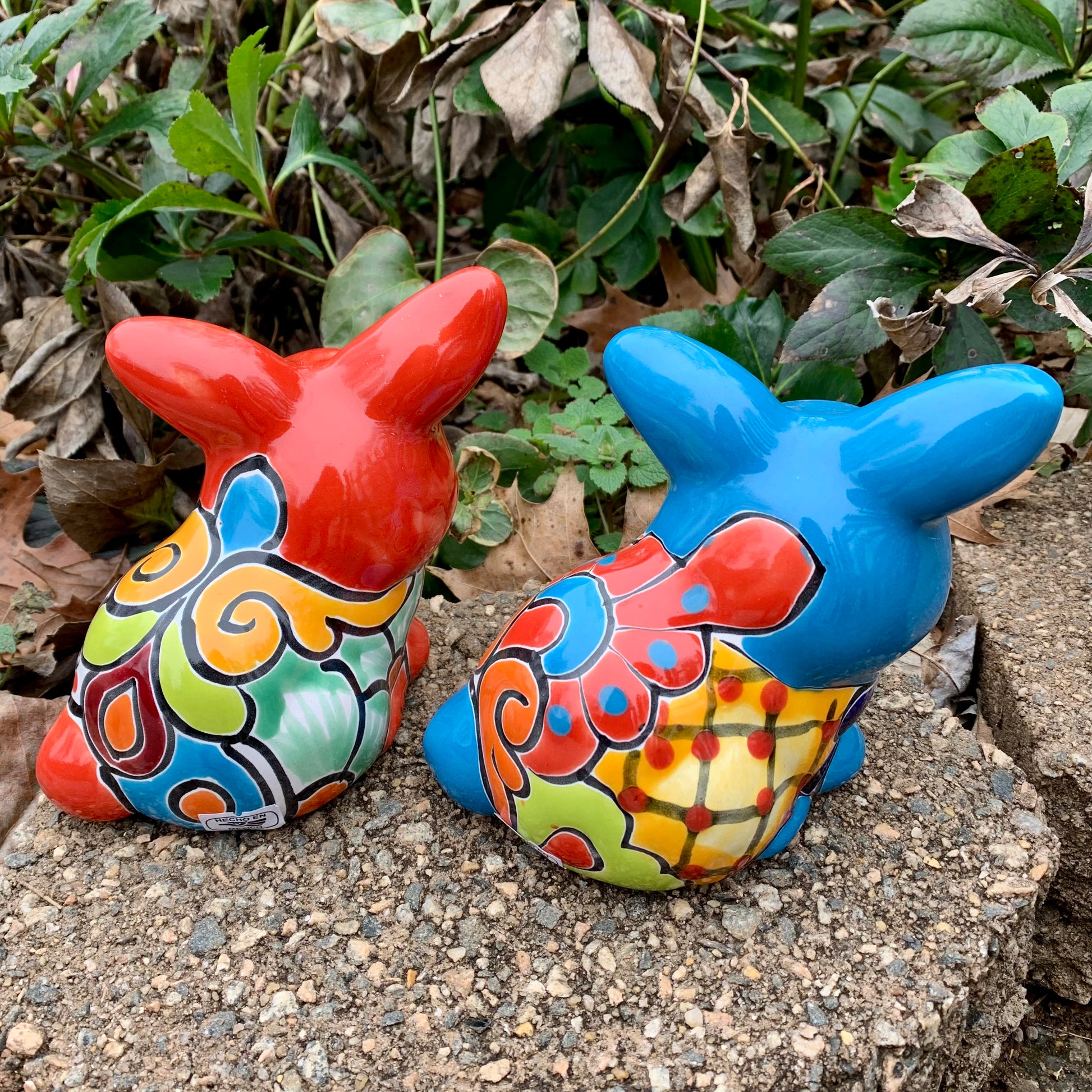 Two colorful Mexican Talavera rabbit figurines on a stone surface with greenery in the background Red and Turquoise back
