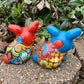 Two colorful Mexican Talavera rabbit figurines on a stone surface with greenery in the background Red and Turquoise back
