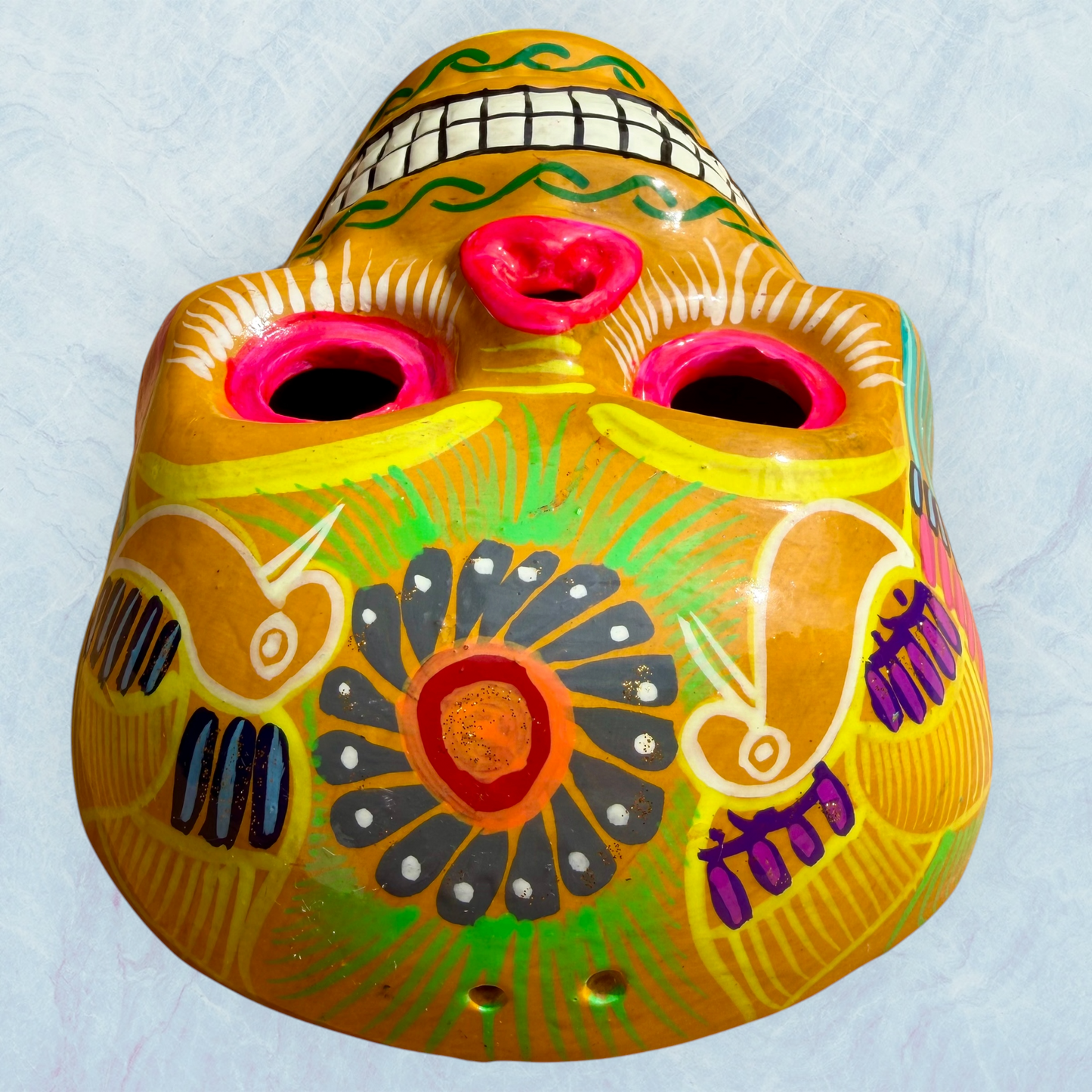 A clay, yellow with floral designs, Mexican-style skeleton skull face mask hanging on a soft blue surface, top showing forehead view.