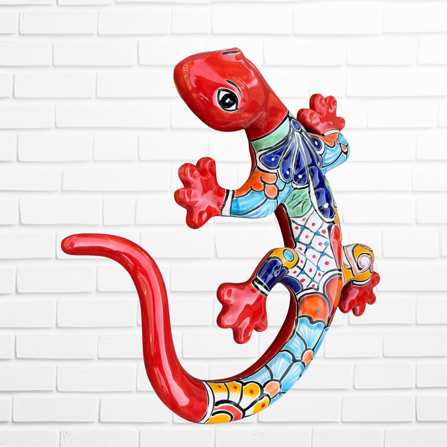 A colorful Talavera ceramic wall salamander lizard art piece with a glossy finish, featuring a red head and tail with a patterned body, displayed against a white brick wall background, left side body view.