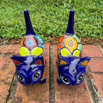 Two colorful hand-painted talavera ceramic cat figurines in cobalt blue with multi color designs standing on a brick path with a green lawn top side view.