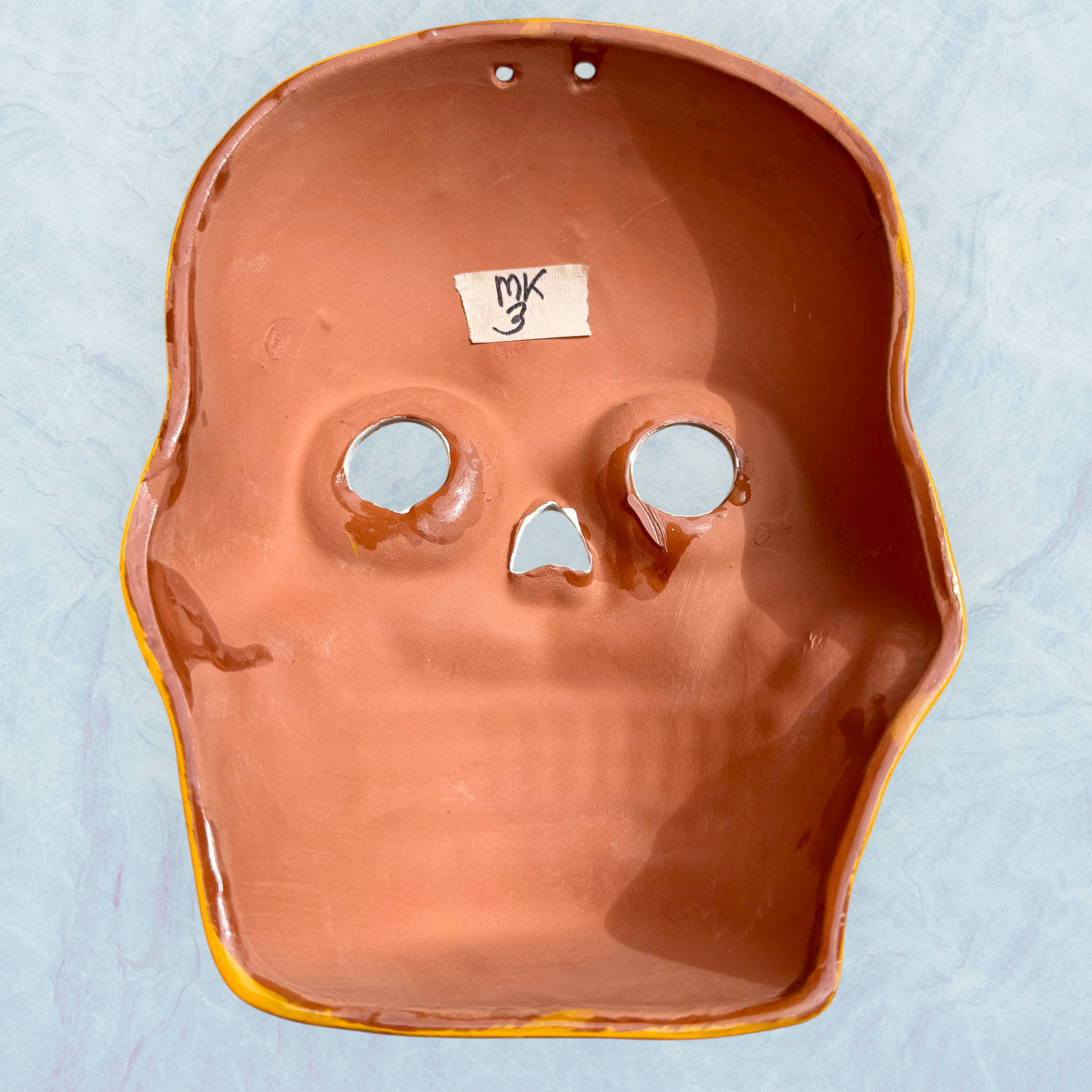 A clay,  golden yellow with floral designs, Mexican-style skeleton skull face mask hanging on a soft blue surface, back view.
