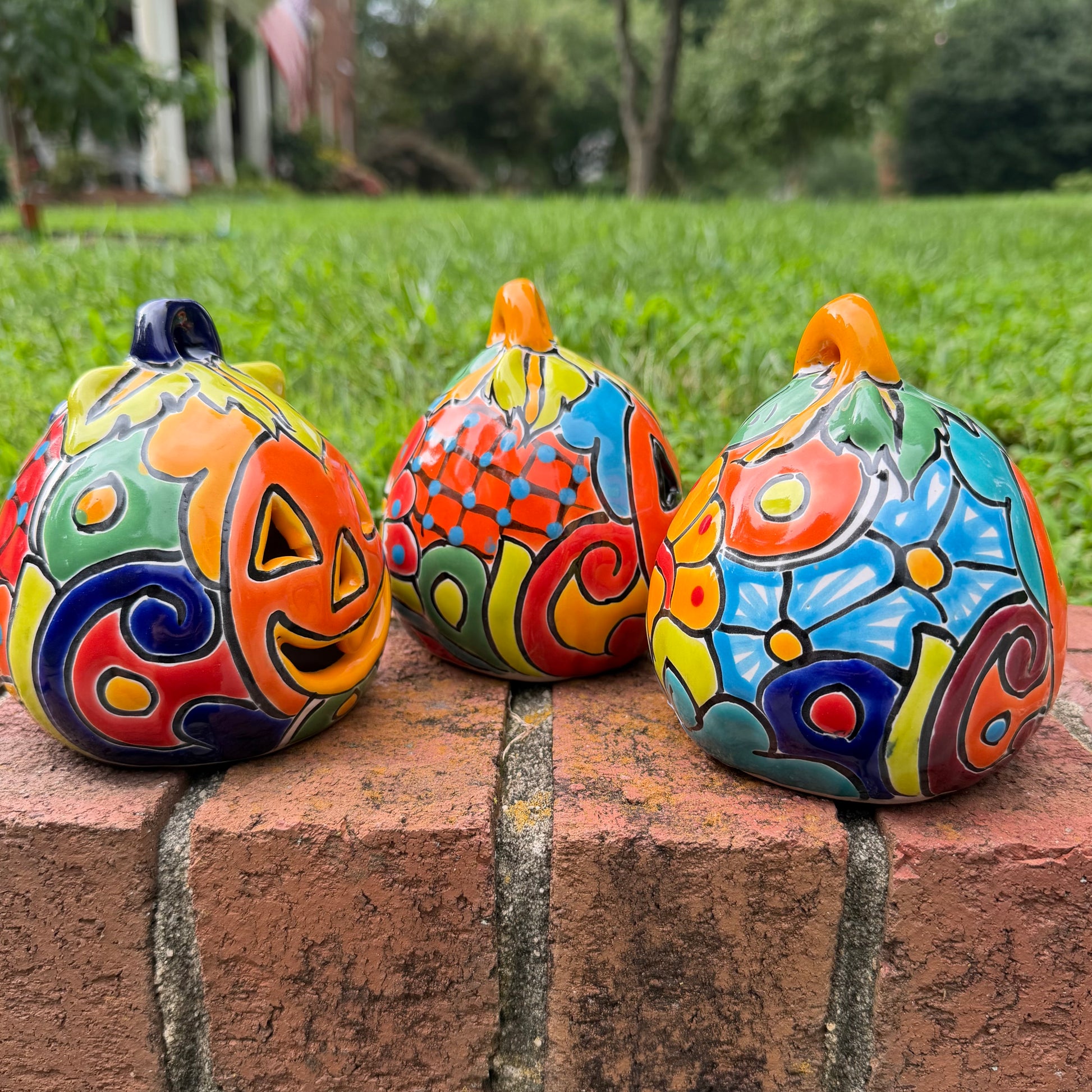 Three hand-painted Talavera ceramic jack-o'-lantern pumpkins with orange faces and yellow, blue, and red green designs, sitting on a brick path in a grassy backyard, left side view.