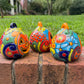 Three hand-painted Talavera ceramic jack-o'-lantern pumpkins with orange faces and yellow, blue, and red green designs, sitting on a brick path in a grassy backyard, left side view.