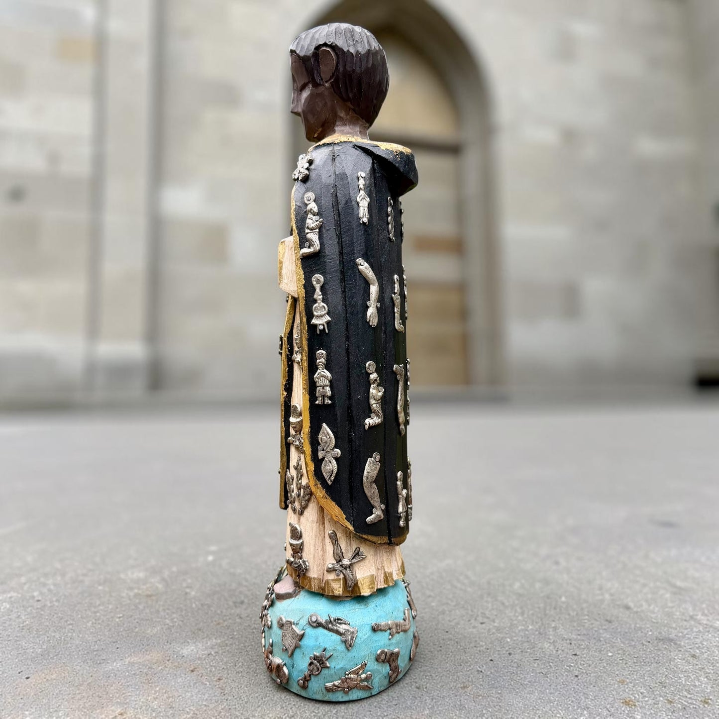 A carved milagros religious figurine of  Saint Martin in ornate black and gold vestments with silver cross details, photographed outdoors in front of a blurred stone archway. Right side view if Saint Martin.