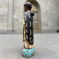 A carved milagros religious figurine of  Saint Martin in ornate black and gold vestments with silver cross details, photographed outdoors in front of a blurred stone archway. Right side view if Saint Martin.