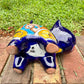 A glossy talavera ceramic cat figurine decorated with vibrant hand-painted patterns in blue, orange, yellow, and red, standing on a brick path with a green lawn and a house in the blurred background. View of the figurines bottom.