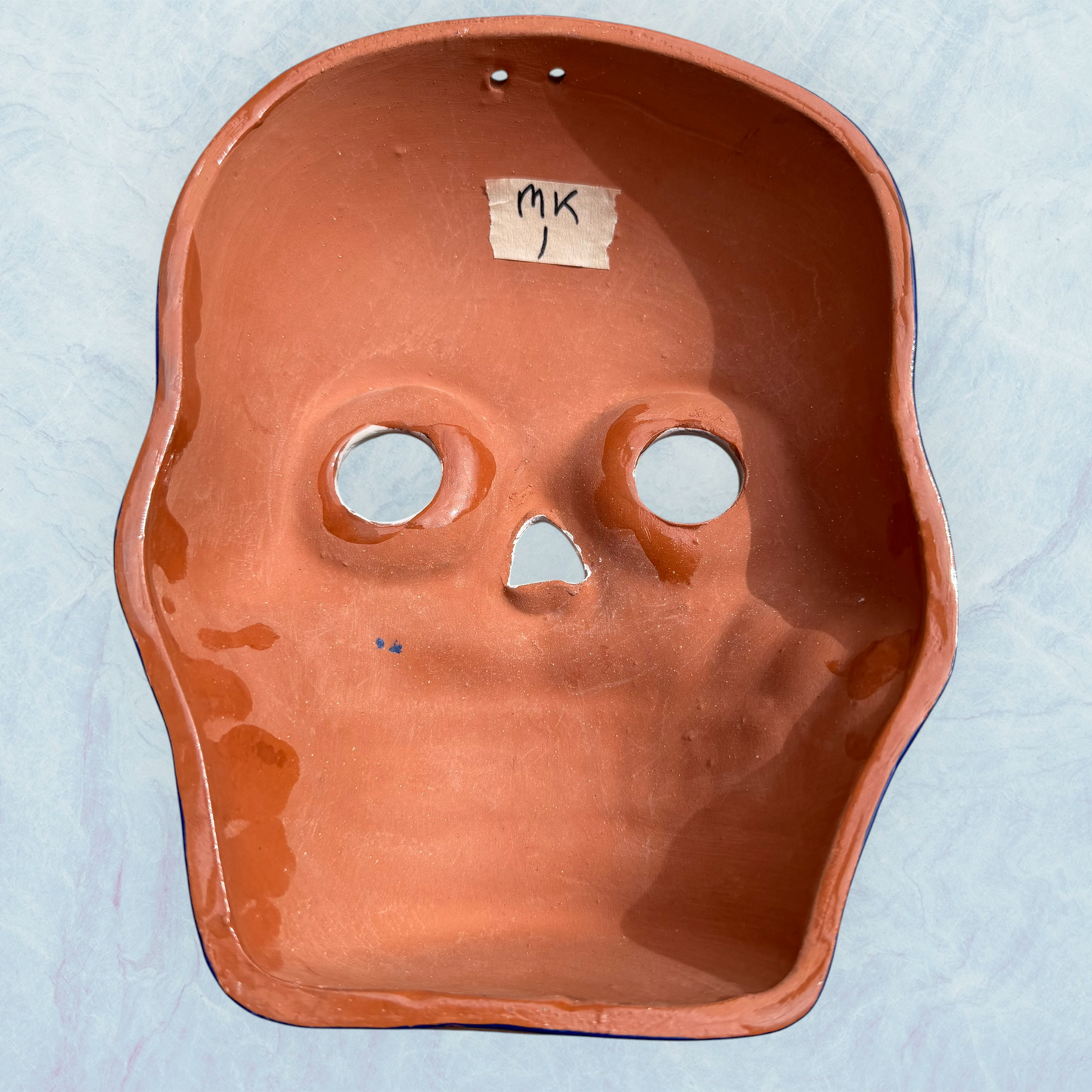 A clay, blue with white designs, Mexican-style skeleton skull face mask hanging on a soft blue surface, back view.