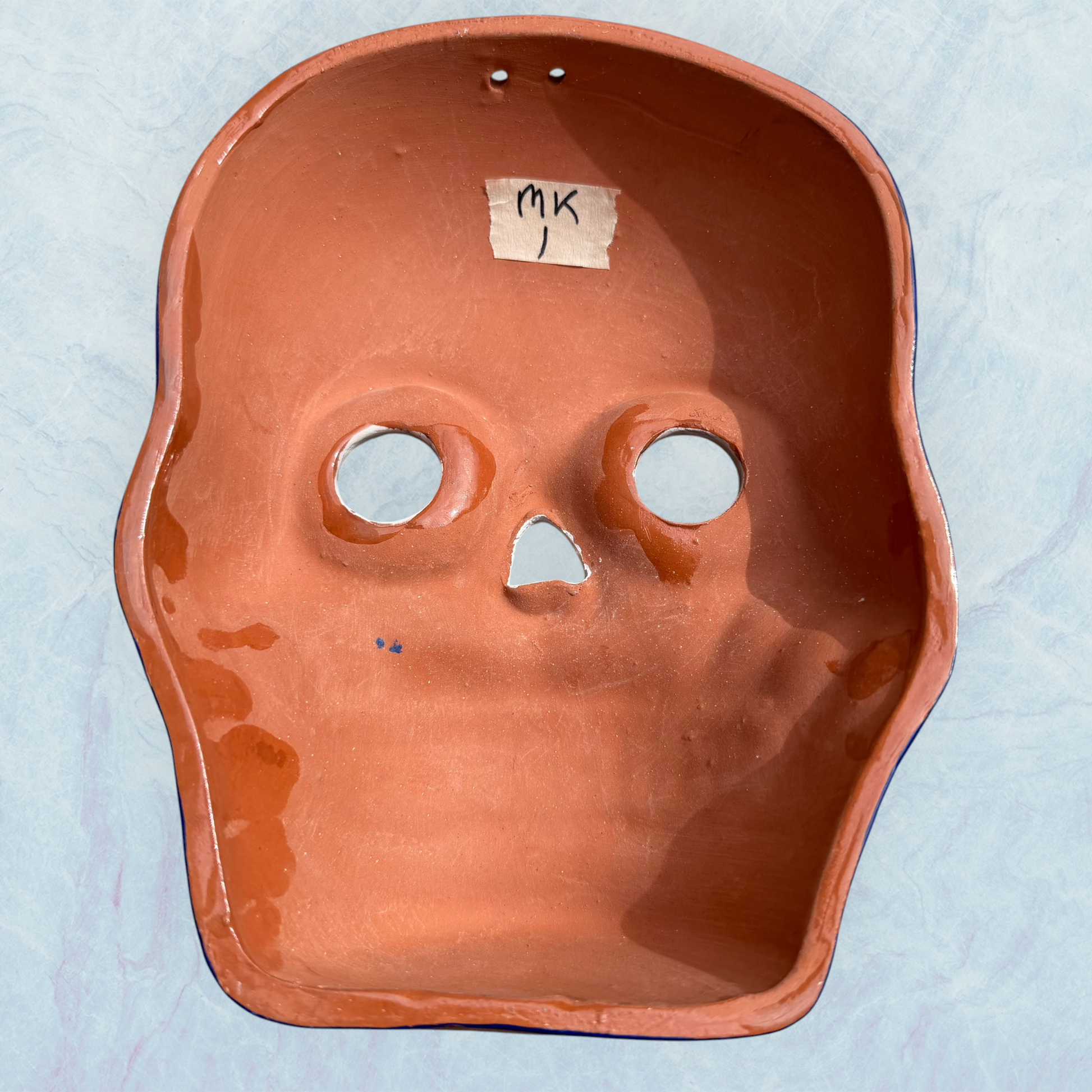 A clay, blue with white designs, Mexican-style skeleton skull face mask hanging on a soft blue surface, back view.