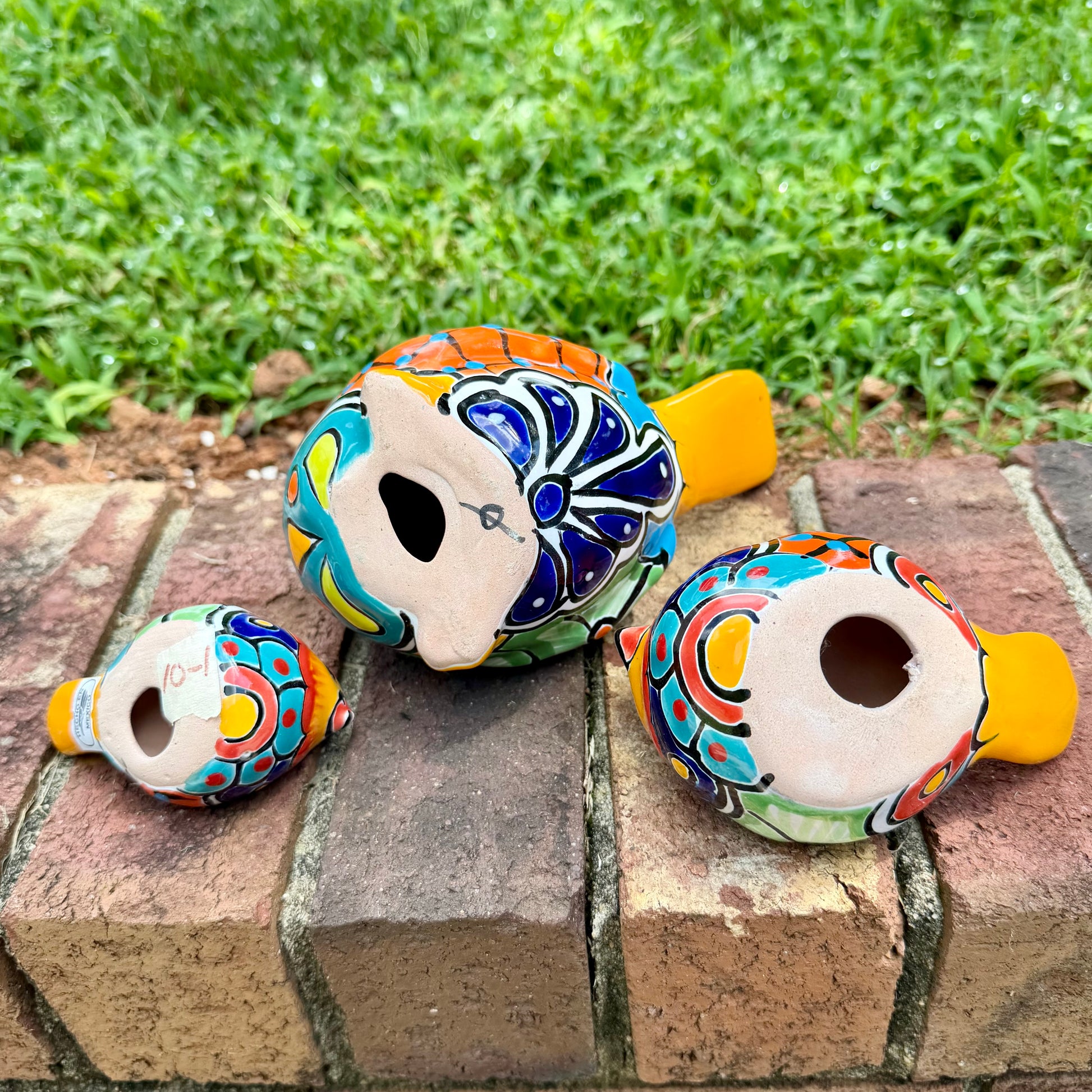 Three hand-painted ceramic bird figurines in yellow, orange  blue, red, and black, displayed on a brick surface with a green grass background bottom view.