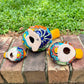 Three hand-painted ceramic bird figurines in yellow, orange  blue, red, and black, displayed on a brick surface with a green grass background bottom view.