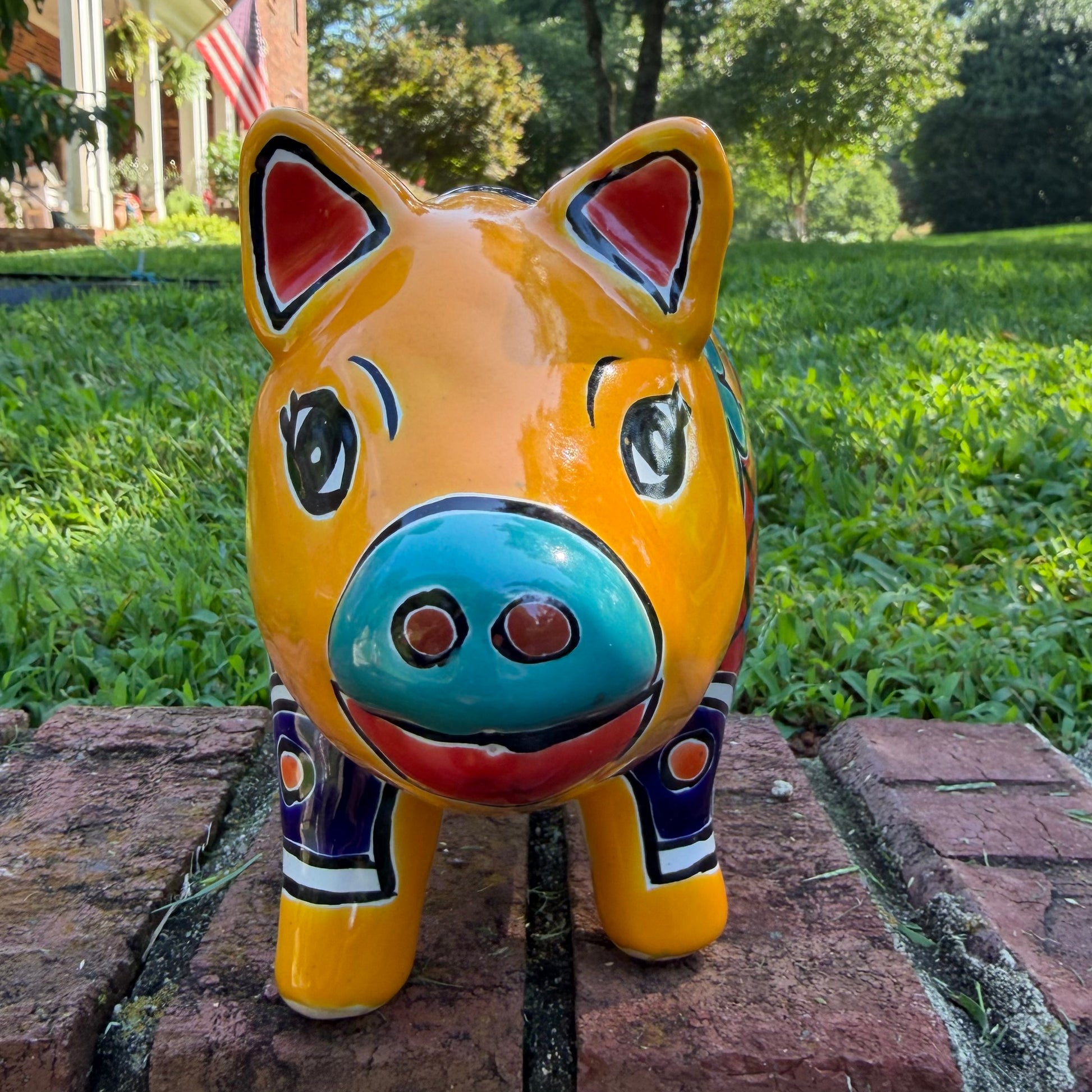 A colorful ceramic pig planter with a yellow-orange face and body, decorated with blue, green, red, and white floral patterns, sitting on a brick surface with a blurred outdoor garden background. Front face close-up of the pig pot.