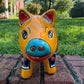 A colorful ceramic pig planter with a yellow-orange face and body, decorated with blue, green, red, and white floral patterns, sitting on a brick surface with a blurred outdoor garden background. Front face close-up of the pig pot.