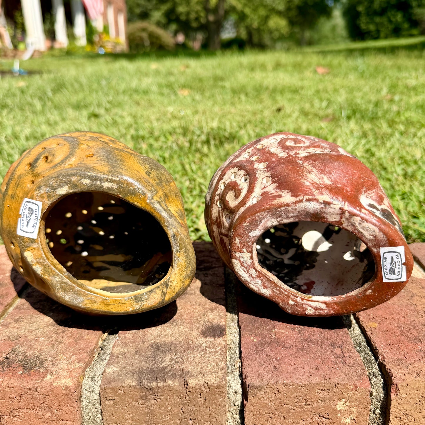 Two decorative clay sugar skull ornaments with Day of the Dead style painted patterns, displayed side-by-side on a brick surface outdoors with a grassy lawn and trees in the background, bottom of skull.