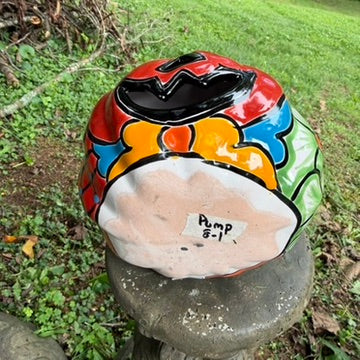 A hand-painted talavera halloween  jack-o'-lantern candle holder in red, green, black, and orange, displayed on a stone pedestal in an outdoor garden setting bottom of pumpkin view.