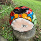 A hand-painted talavera halloween  jack-o'-lantern candle holder in red, green, black, and orange, displayed on a stone pedestal in an outdoor garden setting bottom of pumpkin view.
