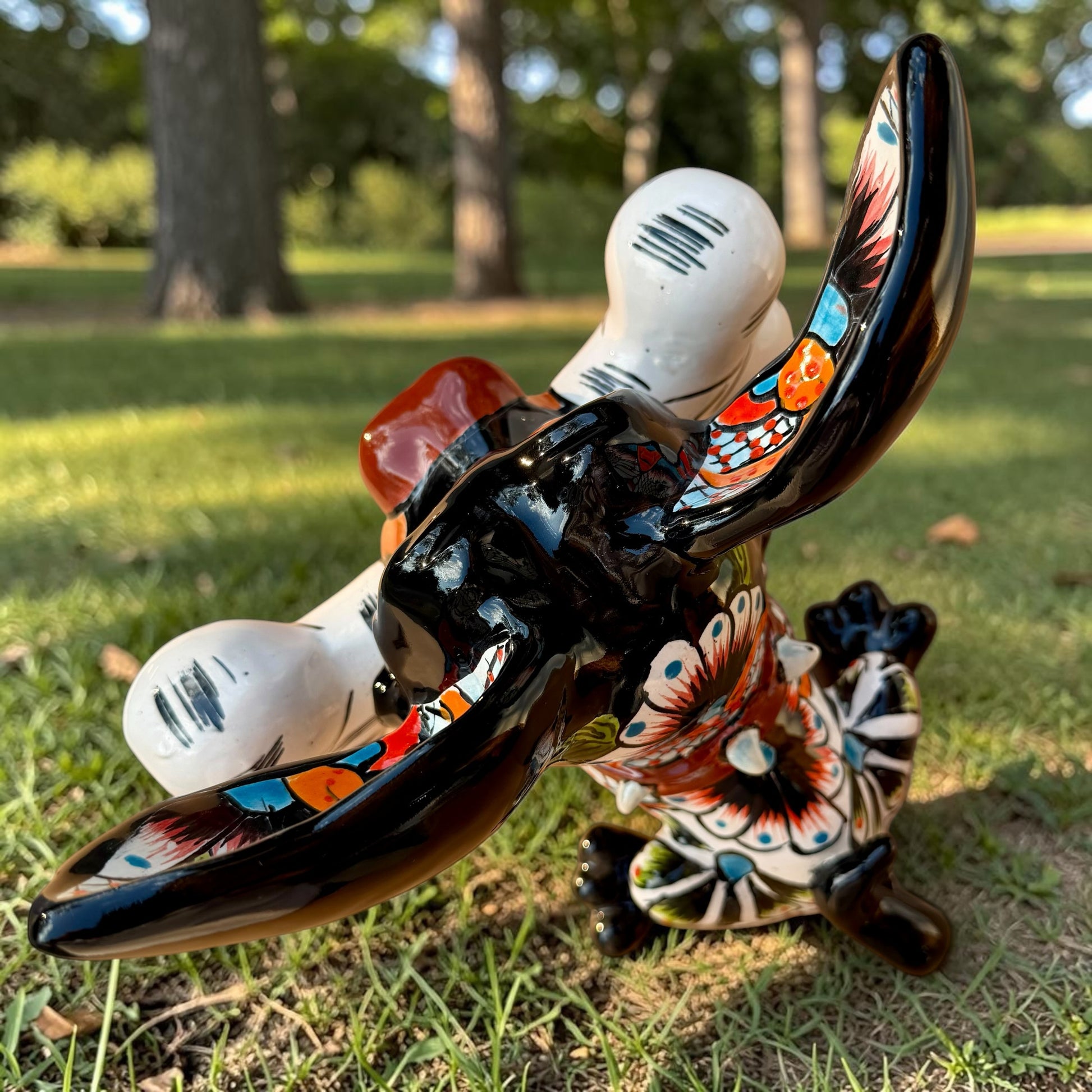 A colorful Talavera dog statue holding a big white bone, photographed outdoors on grass with trees in the background. The dog has a glossy black body, blue eyes, an orange snout, and multicolored patterned ears.  Top view