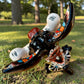 A colorful Talavera dog statue holding a big white bone, photographed outdoors on grass with trees in the background. The dog has a glossy black body, blue eyes, an orange snout, and multicolored patterned ears.  Top view