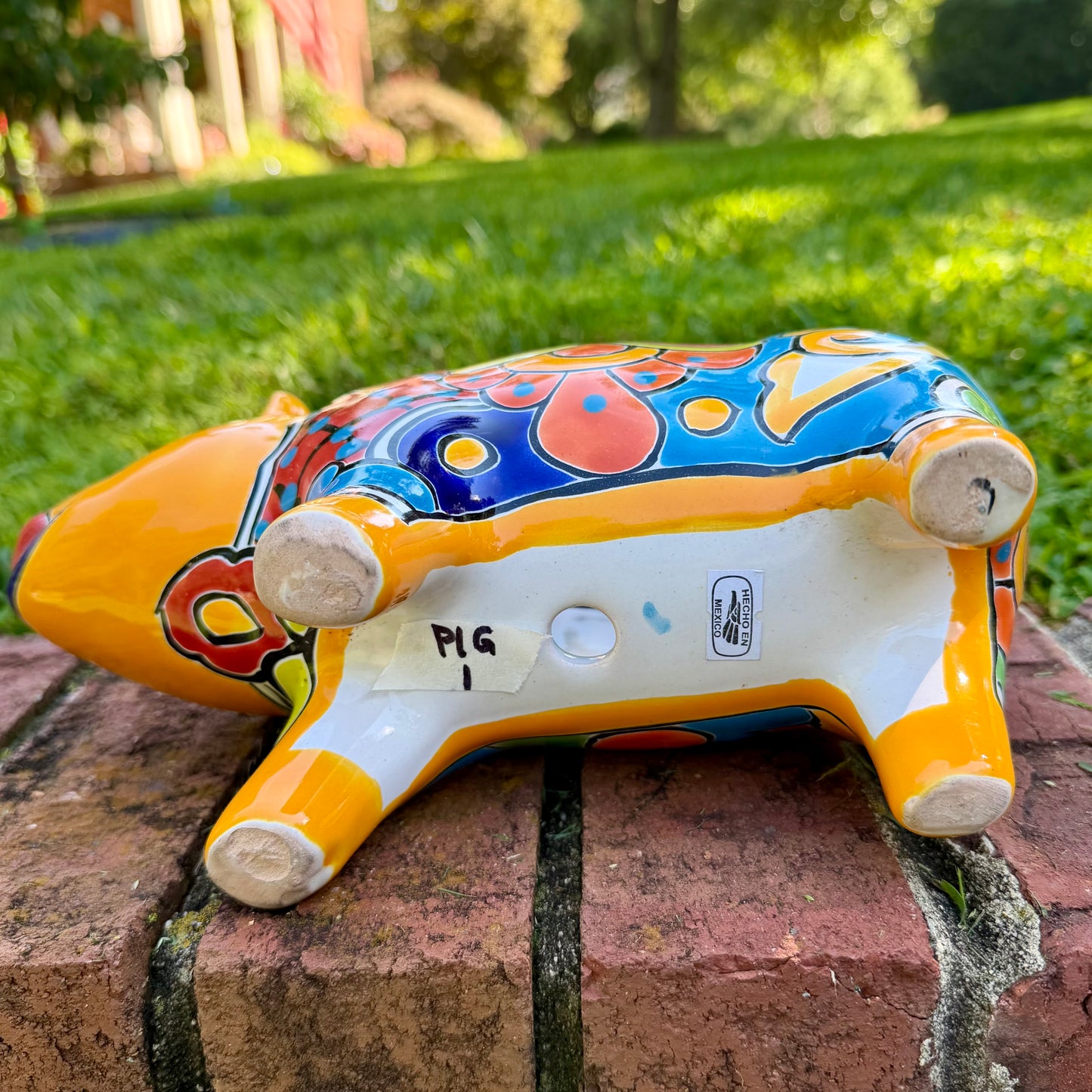 A colorful hand-painted yellow Talavera pig figurine standing on a brick surface with a blurred outdoor garden background, bottm drain hole.