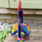 A colorful hand-painted ceramic rooster figurine standing on a rustic wooden surface in front of a green wooden door, back tail view.