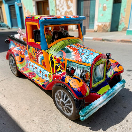 A Mexican Talavera pick up truck decorated with vibrant, colorful  folk art patterns, parked on a street in front of rustic buildings. The truck features a detailed cab, skeleton passengers and a bed carrying farm animals. 