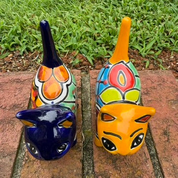 Two colorful talavera ceramic cat figurines standing on a brick surface outdoors with grass and trees blurred in the background. One cat is dark blue with orange, yellow, and red patterns, and the other is orange with blue, red, yellow, and white patterns. view from the top
