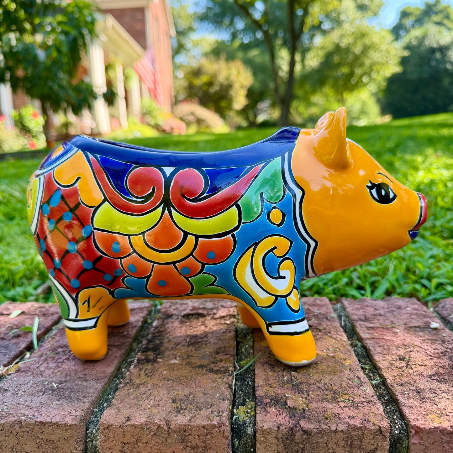 A colorful hand-painted yellow Talavera pig figurine standing on a brick surface with a blurred outdoor garden background, left side view.