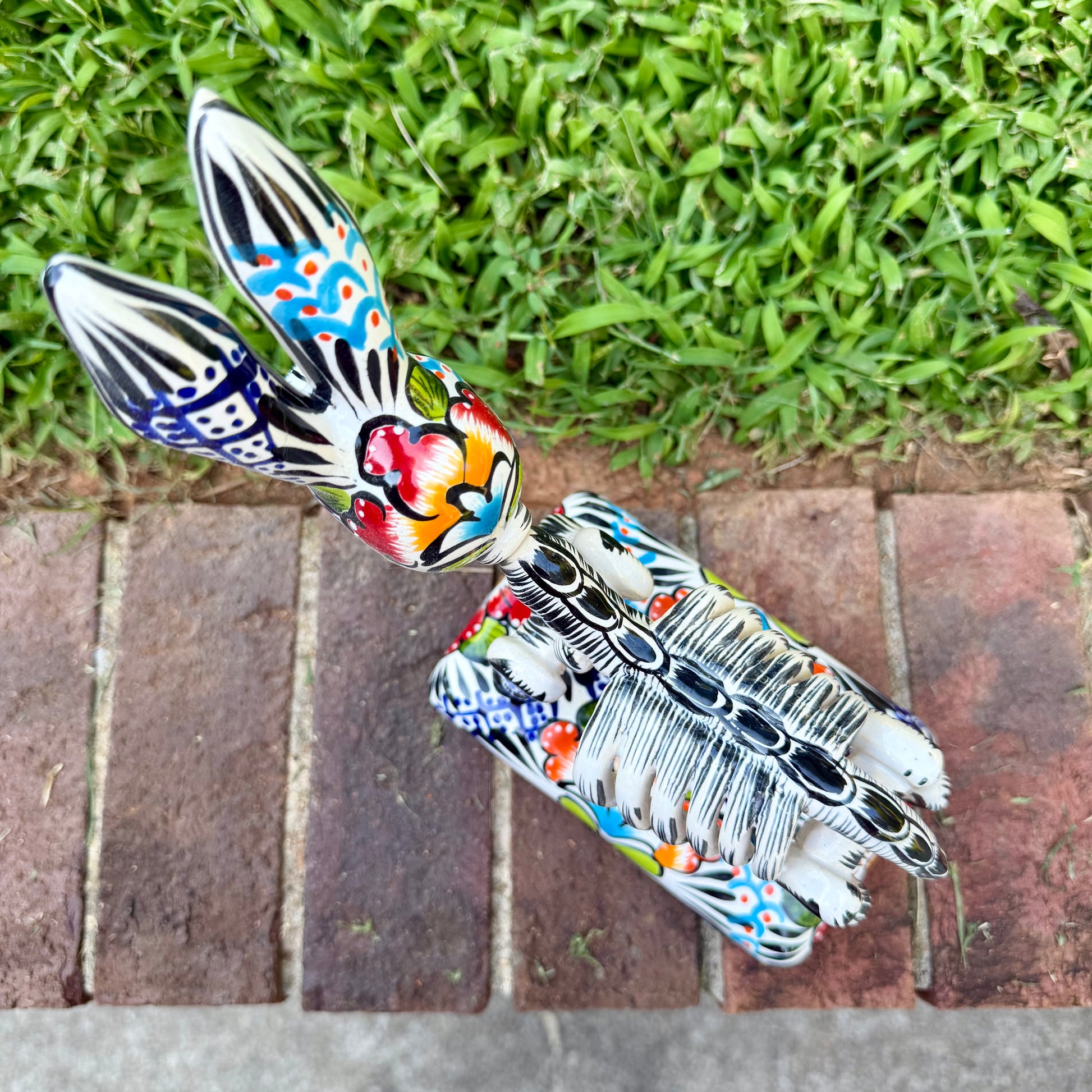 A colorful ceramic donkey Catrina figurine with a skeletal white-and-black design and a floral base, photographed outdoors on a brick surface with a blurred green lawn and house in the background. Top view showing backbone and design on base.