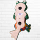 A colorful ceramic frog wall sculpture sitting cross-legged against a white painted brick wall. The frog features a glossy green body with yellow, blue, red, and Back view with hanging holes