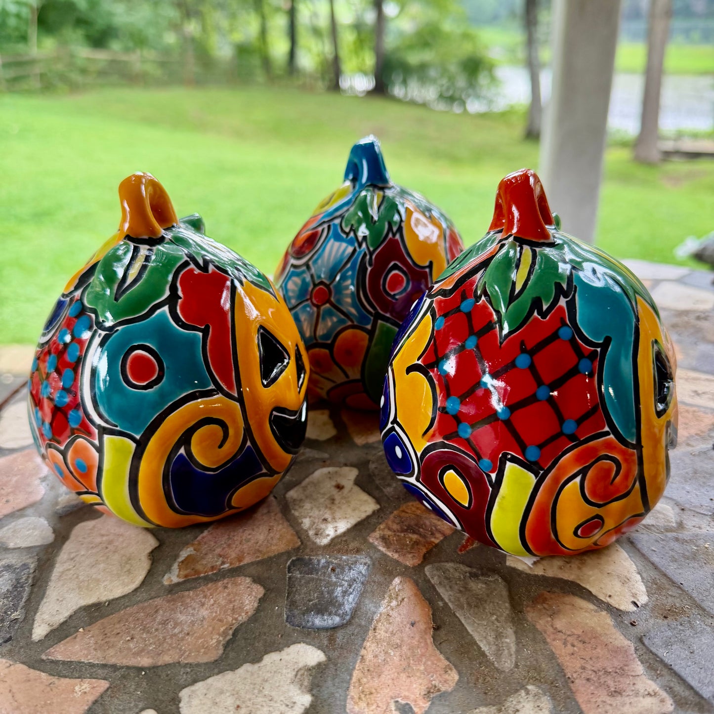 Three colorful hand-painted Talavera jack-o'-lantern pumpkins with multi-color displayed on a stone patio with a blurred green lawn background, right side view.