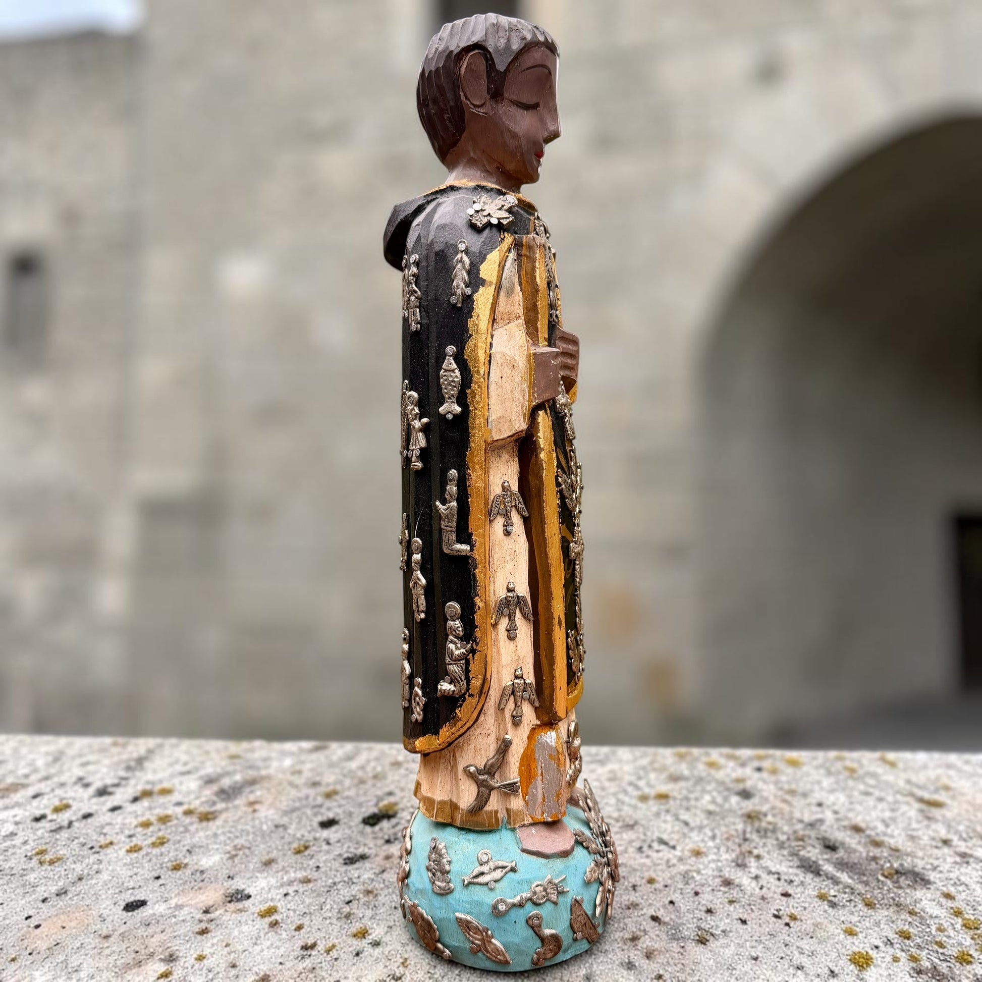 A carved milagros religious figurine of  Saint Martin in ornate black and gold vestments with silver cross details, photographed outdoors in front of a blurred stone archway. Left side view Saint Martin. 