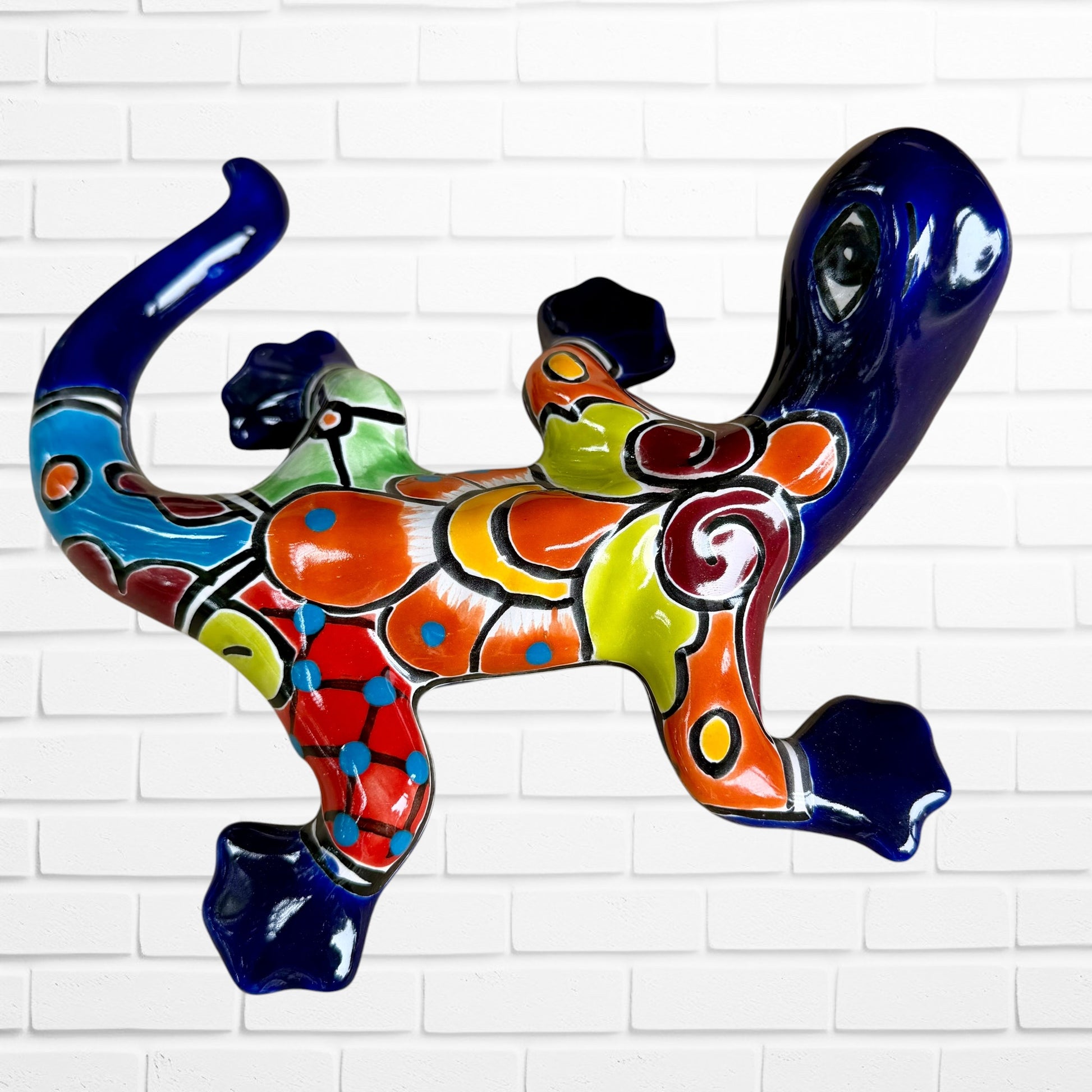 A colorful glazed ceramic lizard wall sculpture with a vibrant multicolor pattern, displayed against a white brick wall background back side leg View.