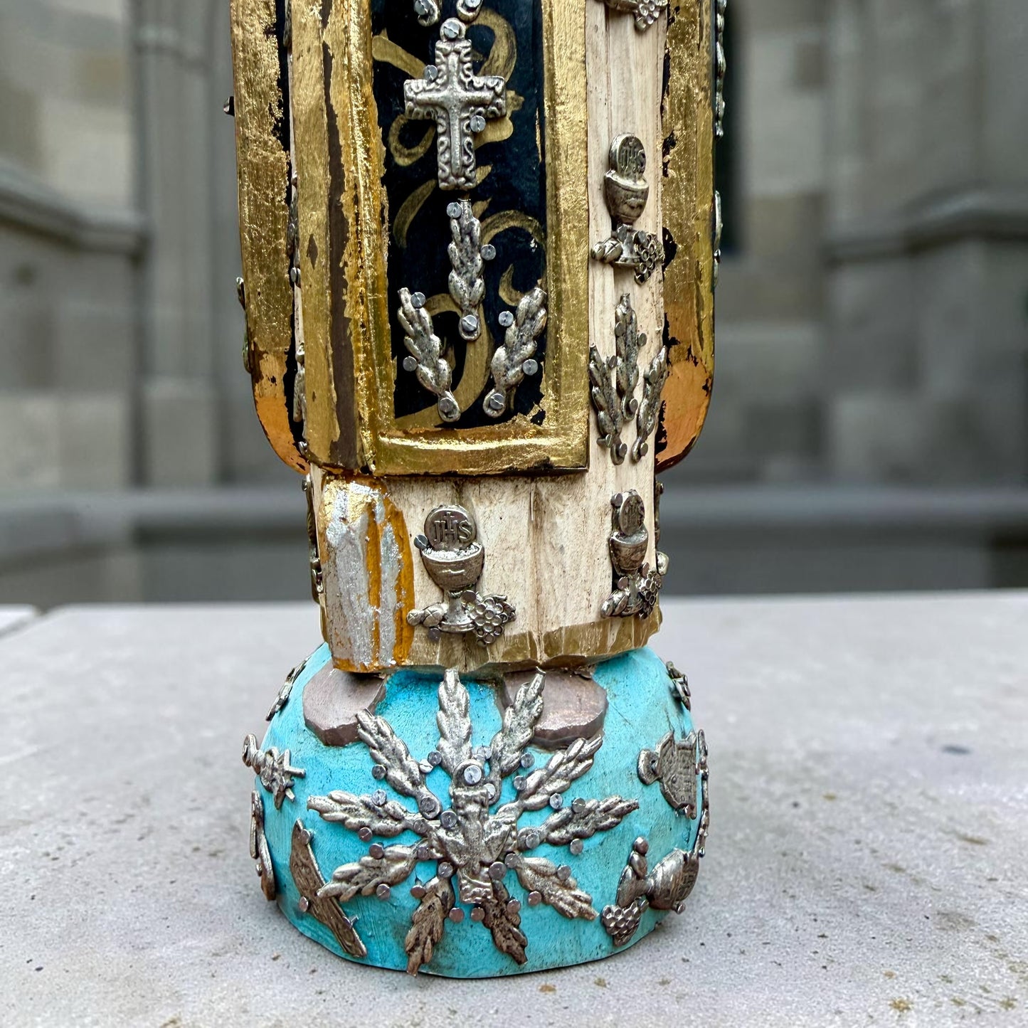A carved milagros religious figurine of  Saint Martin in ornate black and gold vestments with silver cross details, photographed outdoors in front of a blurred stone archway. View of bottom detail.