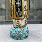A carved milagros religious figurine of  Saint Martin in ornate black and gold vestments with silver cross details, photographed outdoors in front of a blurred stone archway. View of bottom detail.