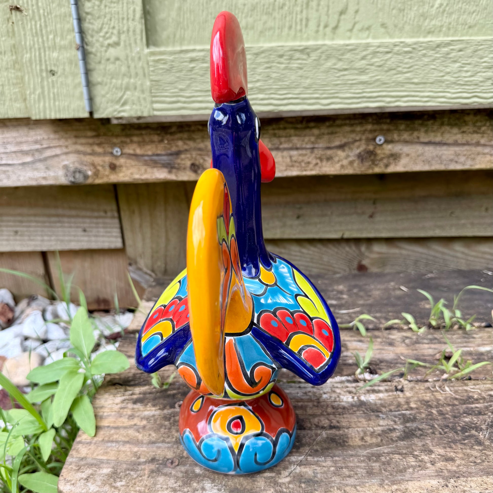 A colorful ceramic rooster figurine standing on a weathered wooden surface outdoors, with a green wooden wall behind it. The rooster is decorated with bright red, yellow, blue, and orange painted details, back of rooster tail view.