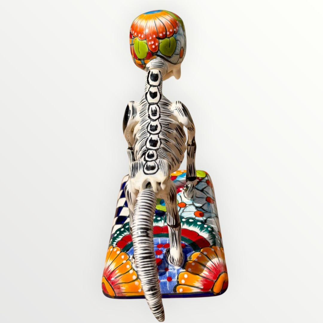 A talavera catrina dog skeleton figurine with colorful hand-painted floral and geometric patterns, standing on a matching decorated base against a plain white background. Back view of dog catrina showing backbone.