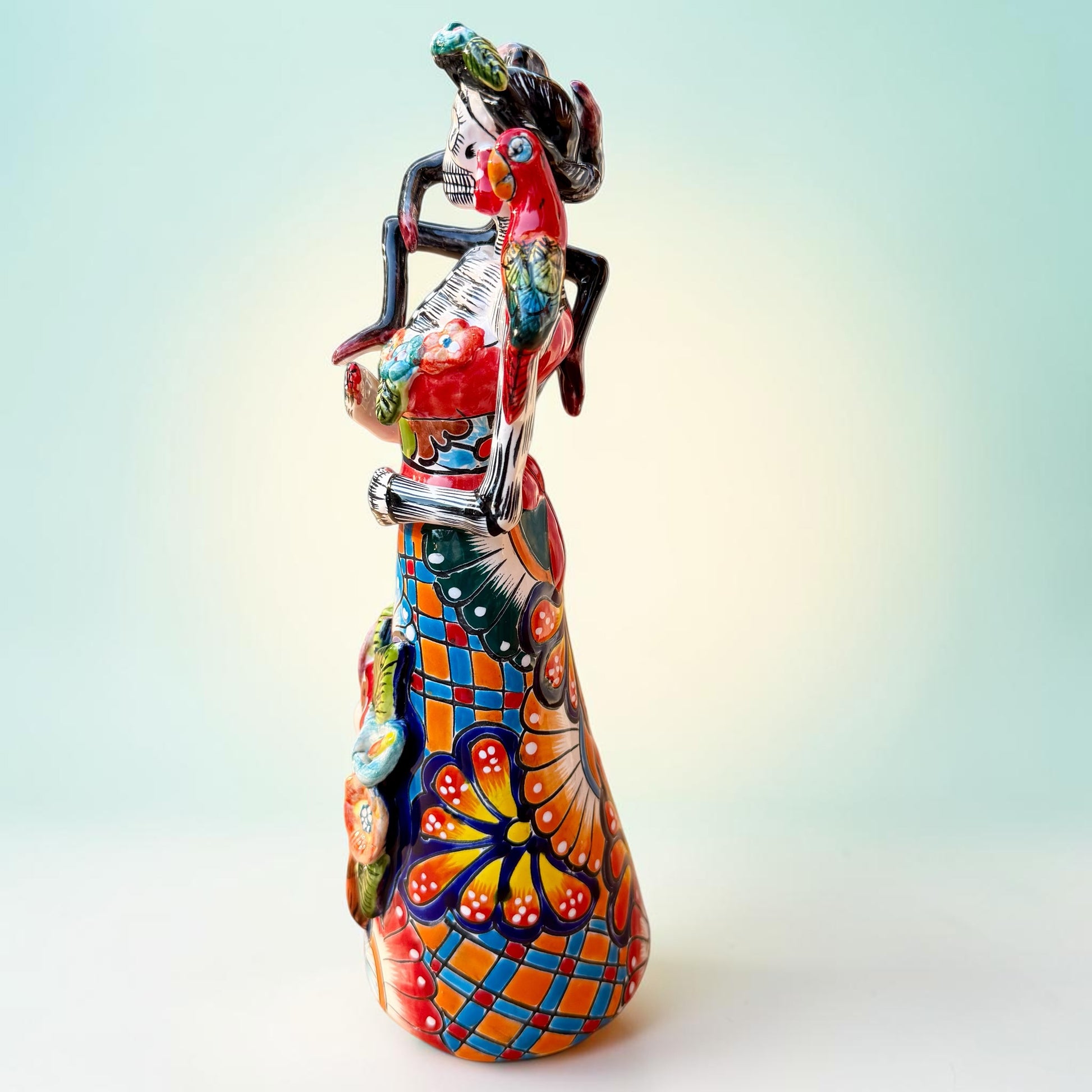 A Talavera Day of the Dead (Día de los Muertos) Frida figurine  with a monkey on her shoulder and a red parrot on her other shoulder, set against a light blue gradient background. Right side view of figurine.