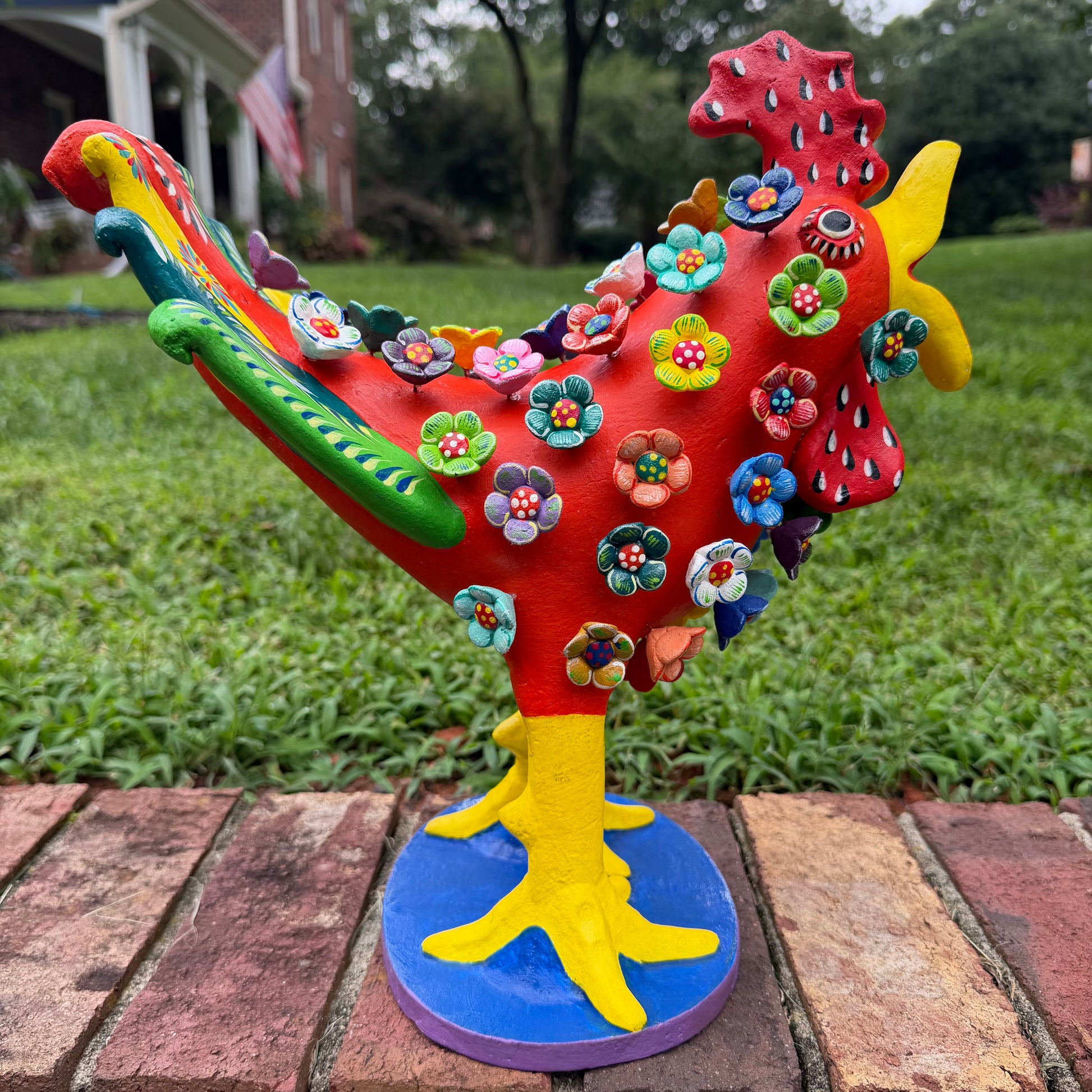 A colorful decorative rooster figurine standing on a brick surface outdoors, left side view, with a grassy lawn and trees in the background. The rooster is primarily red and yellow with many small multicolored flowers and painted details across its body and tail.
