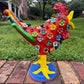 A colorful decorative rooster figurine standing on a brick surface outdoors, left side view, with a grassy lawn and trees in the background. The rooster is primarily red and yellow with many small multicolored flowers and painted details across its body and tail.