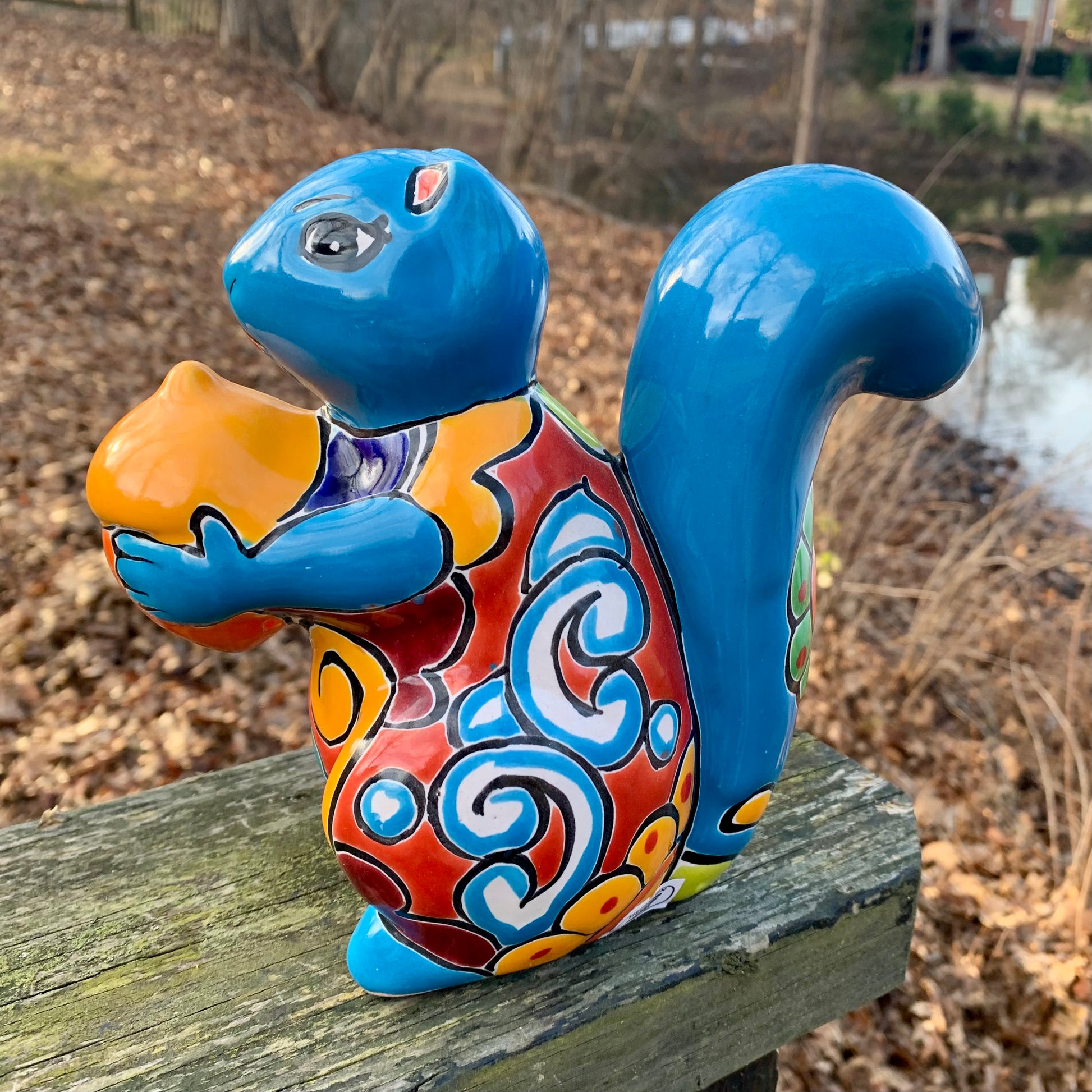 Colorful ceramic squirrel figurine with a blue tail on a wooden surface outdoors.