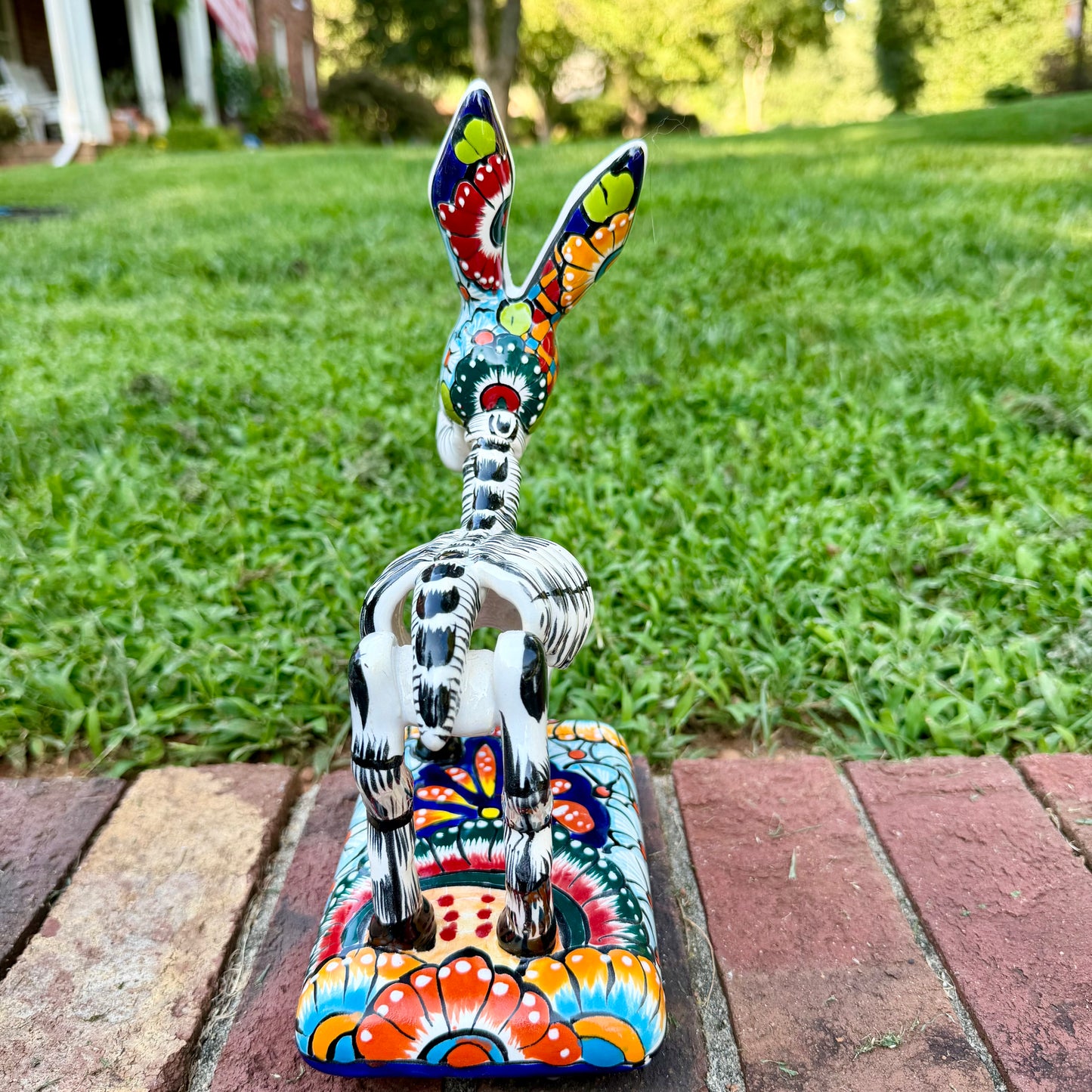 A colorful Catrina donkey figurine standing on a decorated base, photographed outdoors on a brick surface with a grassy yard and house in the background, back view. 