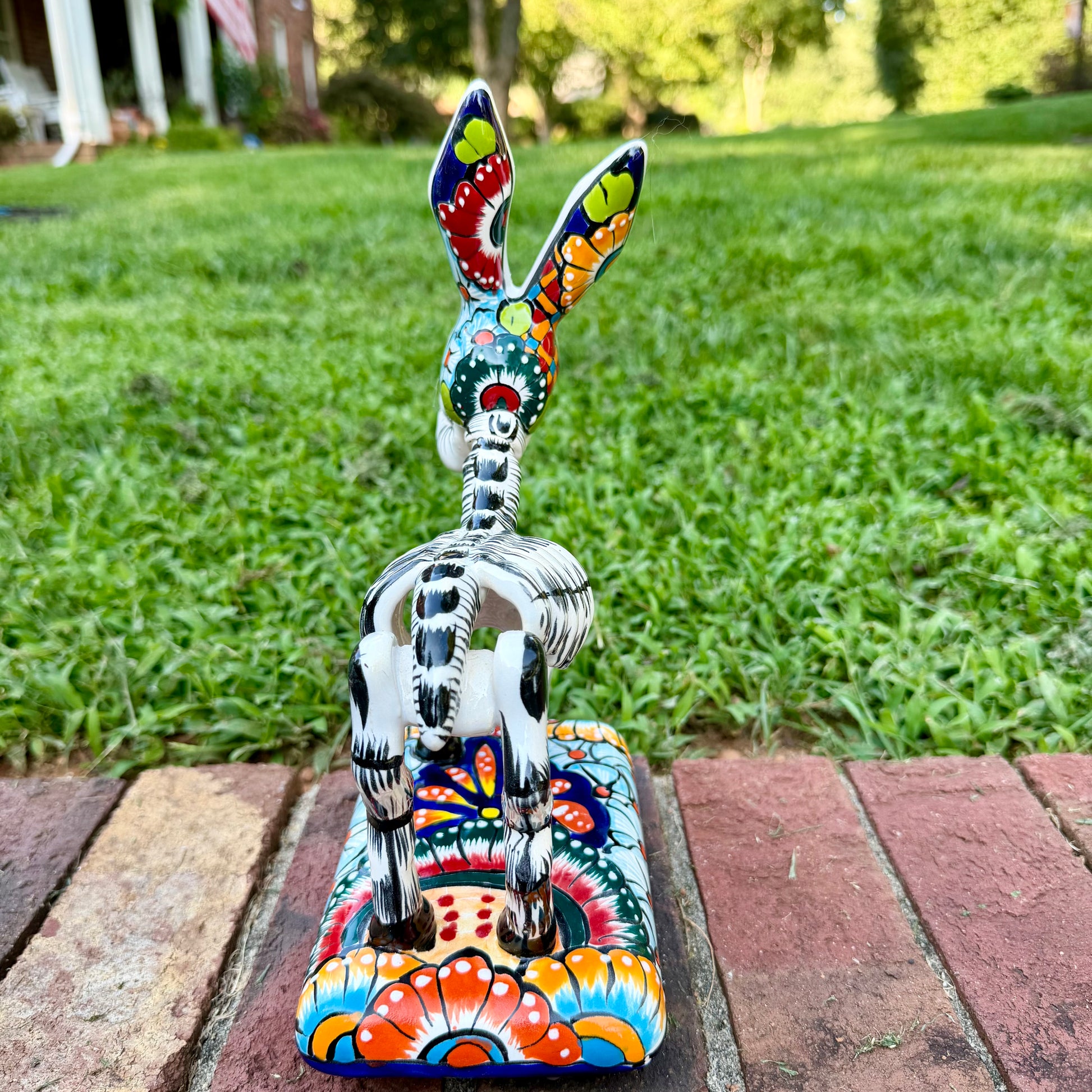 A colorful Catrina donkey figurine standing on a decorated base, photographed outdoors on a brick surface with a grassy yard and house in the background, back view. 