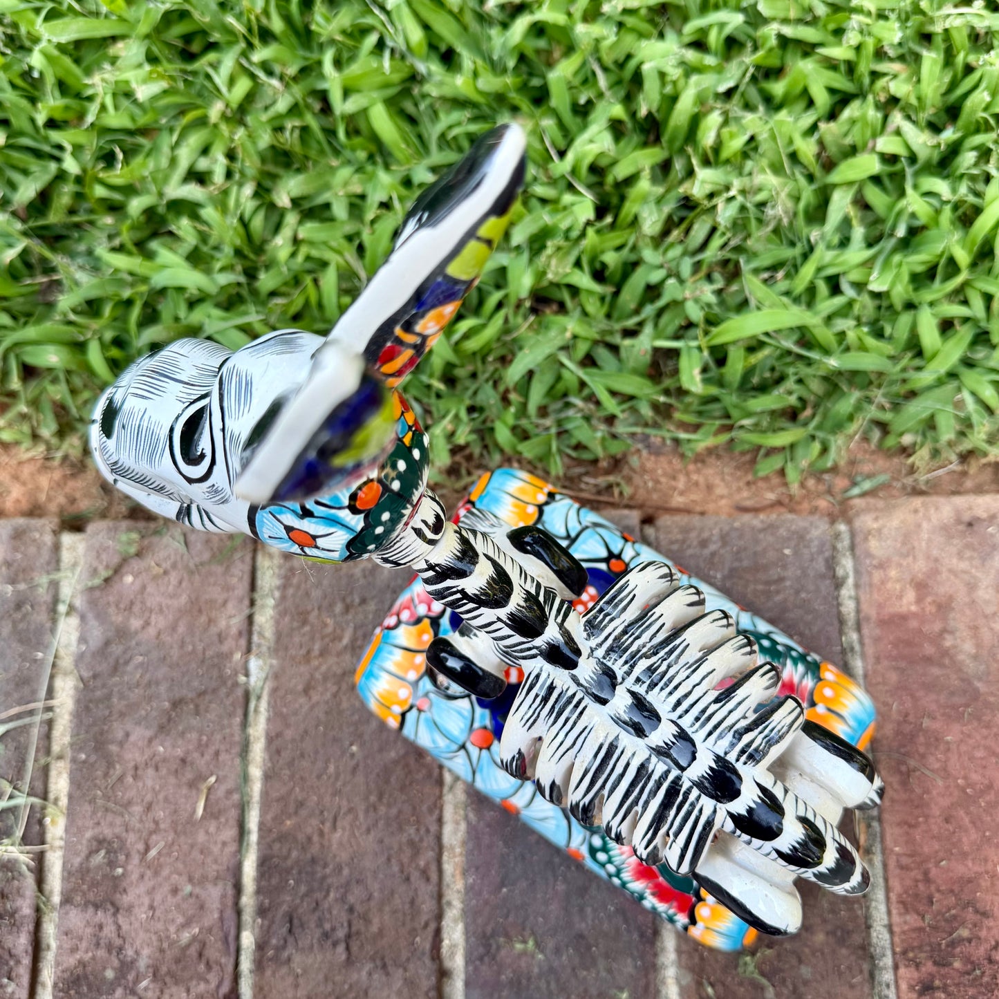 A colorful Catrina donkey figurine standing on a decorated base, photographed outdoors on a brick surface with a grassy yard and house in the background. Top view of donkey showing backbone.