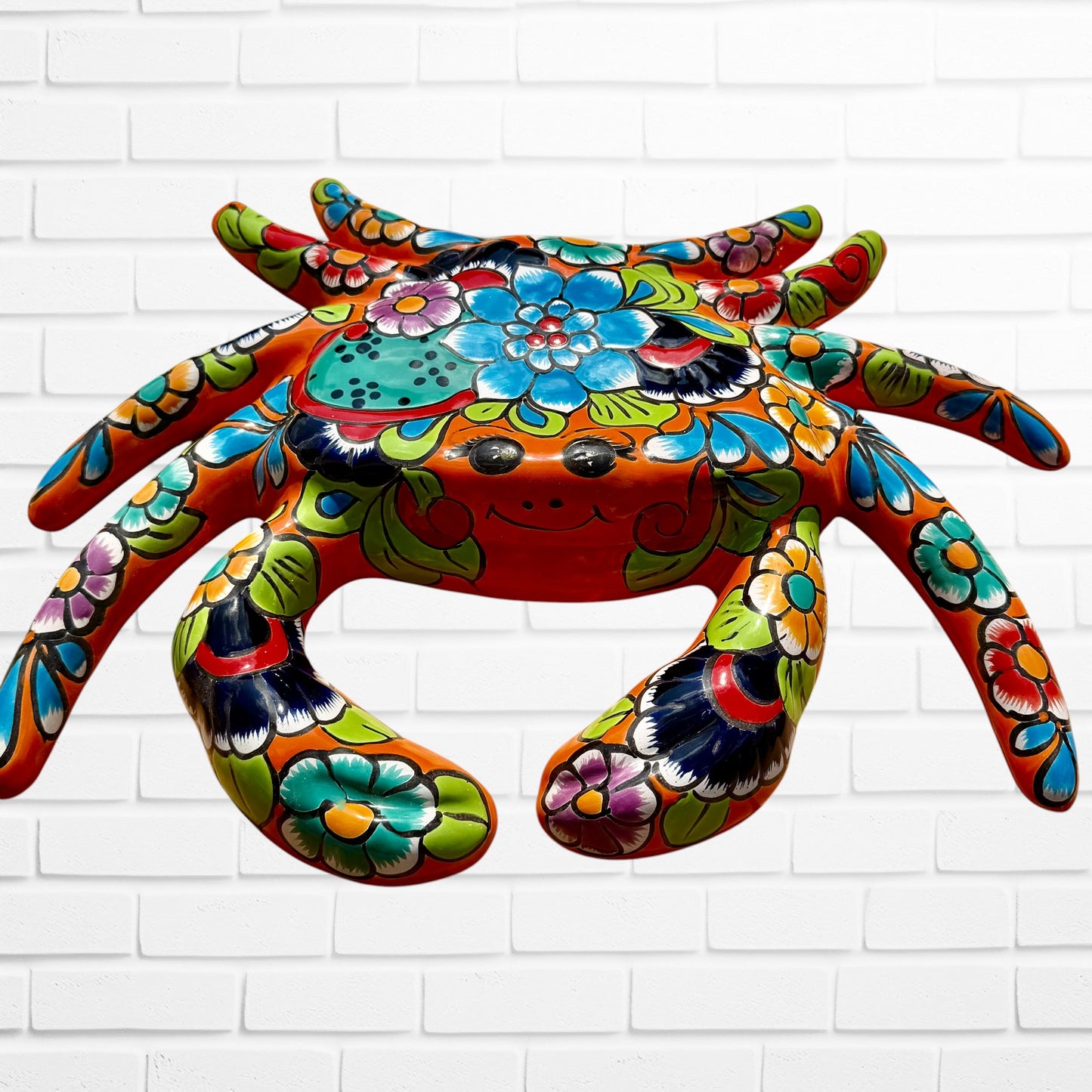A colorful multicolor hand-painted wall art sculpture of a crab, decorated with vibrant floral patterns, shown against a white brick wall background, front face.
