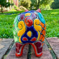 A colorful hand-painted red Talavera ceramic pig figurine standing on a brick surface with a blurred green lawn and trees in the background, back view.
