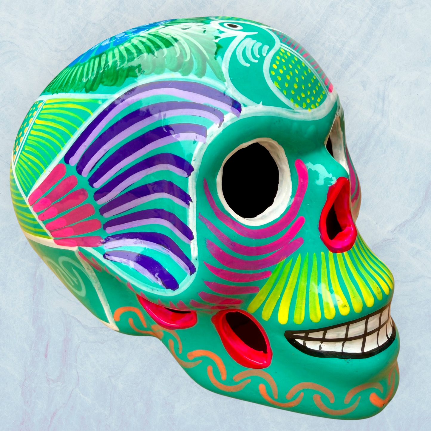 A decorative skull sculpture with intricate colorful sugar skull style patterns painted on a teal colored base, set against a light blue textured background.