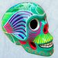 A decorative skull sculpture with intricate colorful sugar skull style patterns painted on a teal colored base, set against a light blue textured background.