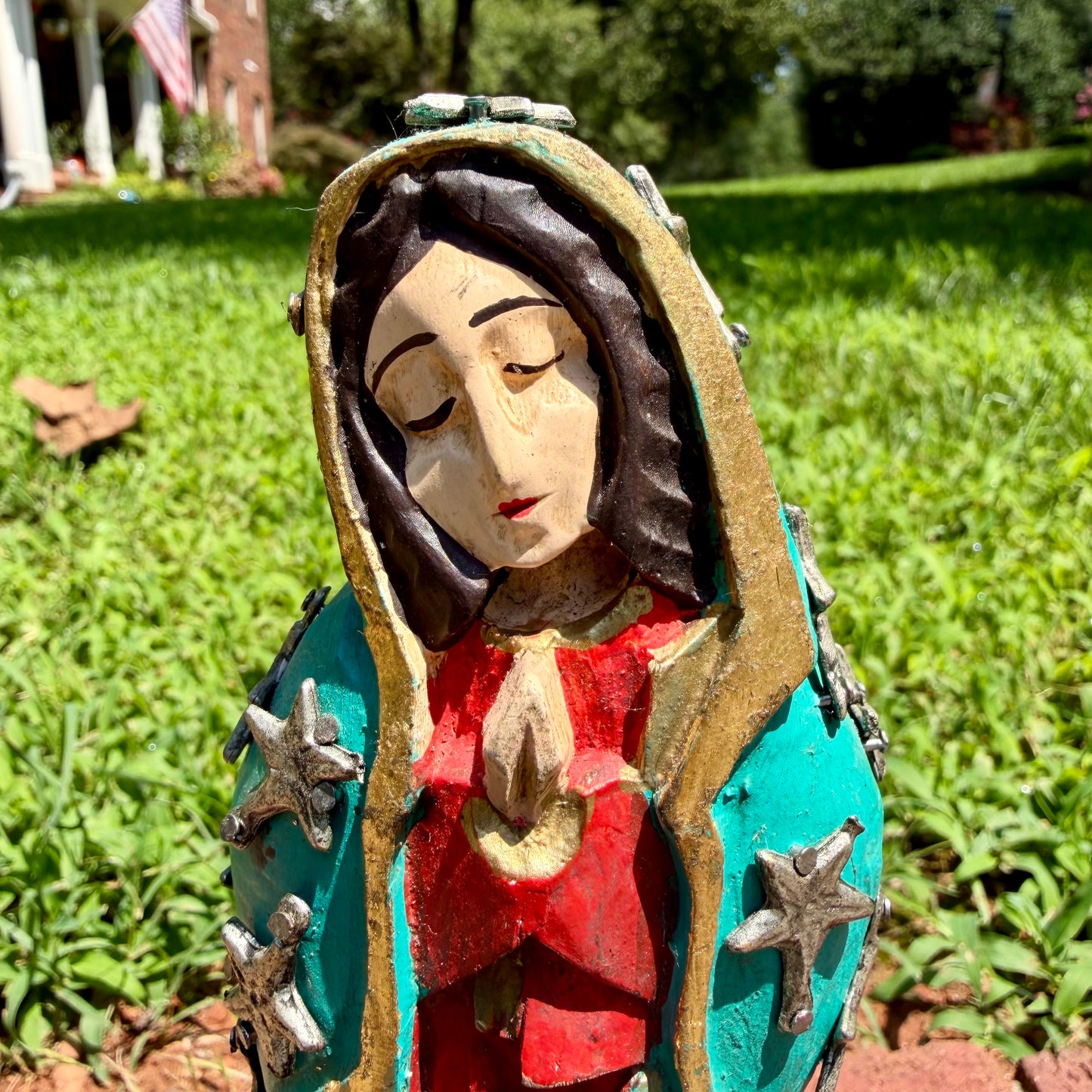 A colorful Milagros Guadalupe  (Virgin Mary) standing on a brick surface outdoors with a grassy yard in the background, close-up of face.